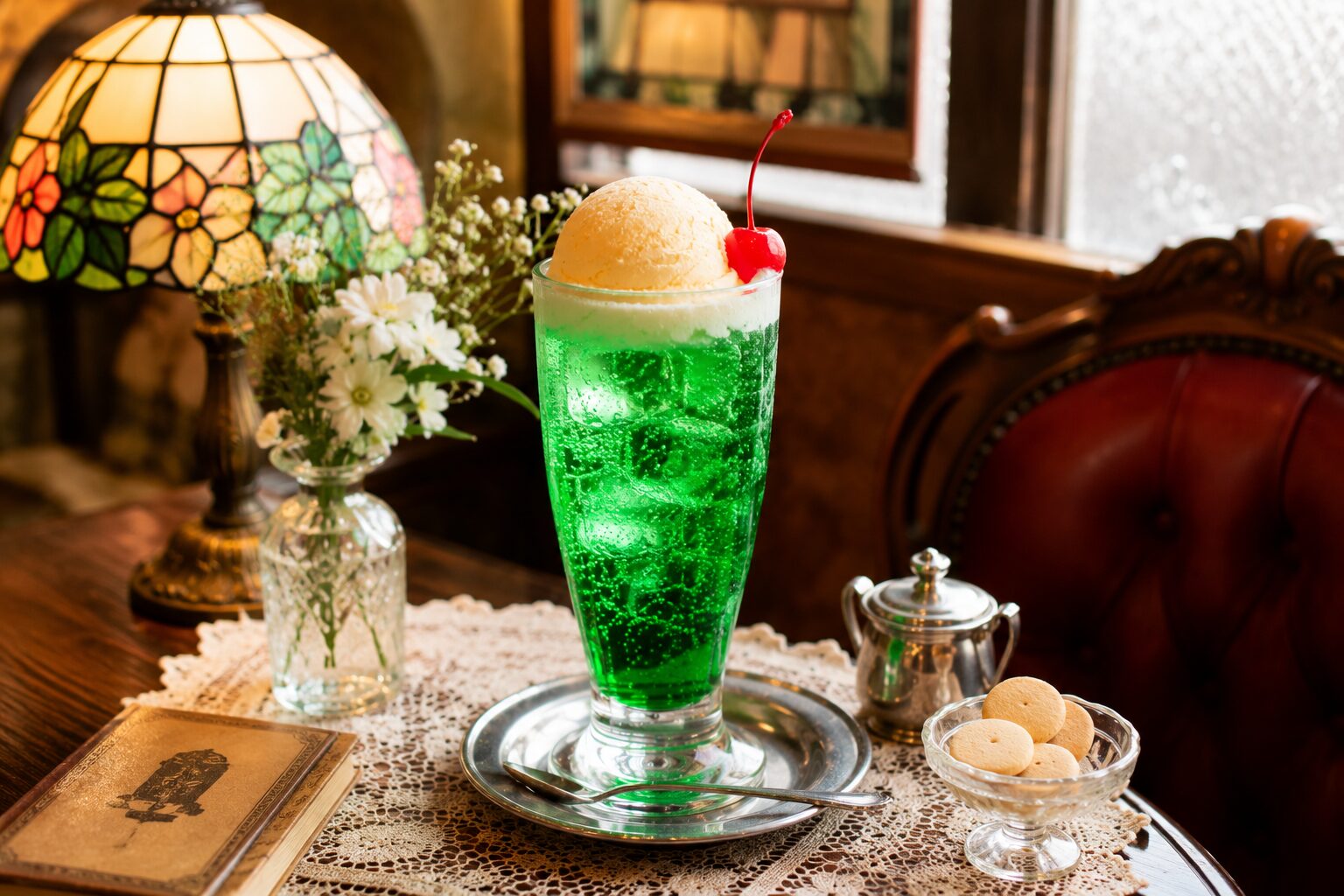 レトロ喫茶の木製テーブルに置かれた鮮やかな緑色のクリームソーダ。バニラアイスとさくらんぼが乗った王道スタイルで、ステンドグラスランプや花瓶に囲まれた温かみのある純喫茶風の空間。